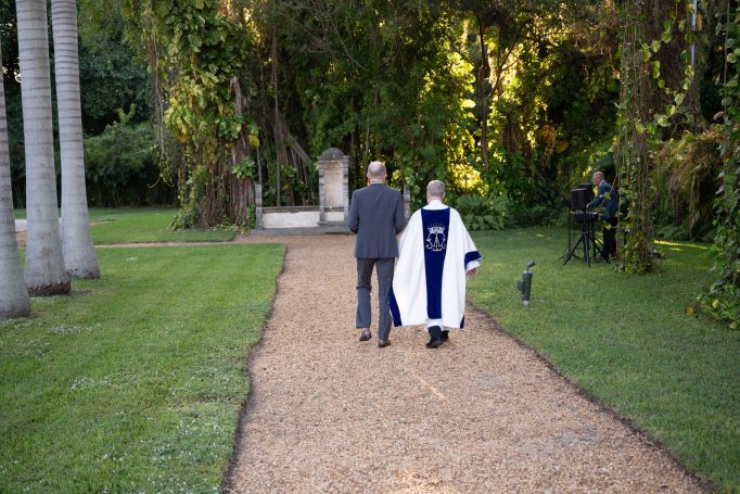Two men walk down a gravel path surrounded by greenery, one in a graduation gown.