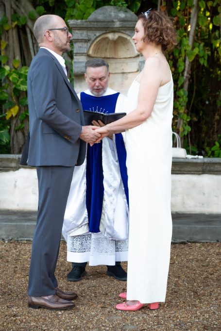 A couple stands together exchanging vows, with an officiant behind them in a garden setting.