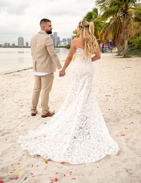 A couple in wedding attire holds hands on a beach with city skyline in the background.