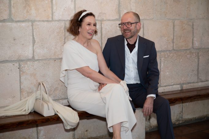 A smiling couple sitting on a bench, dressed elegantly, against a stone wall.