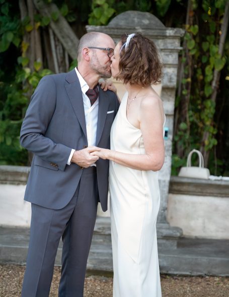 Couple sharing a kiss in elegant wedding attire, surrounded by greenery.