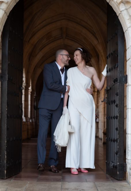 A couple stands by large wooden doors, smiling; one wears a white outfit and the other a suit.