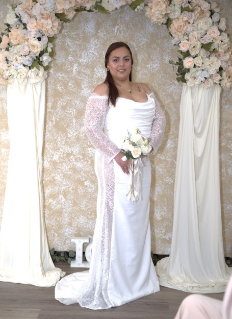 Woman in a white wedding dress holding a bouquet, standing under a floral arch.