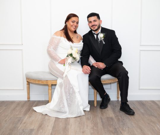 Bride in a white gown and groom in a black suit seated together, both smiling.