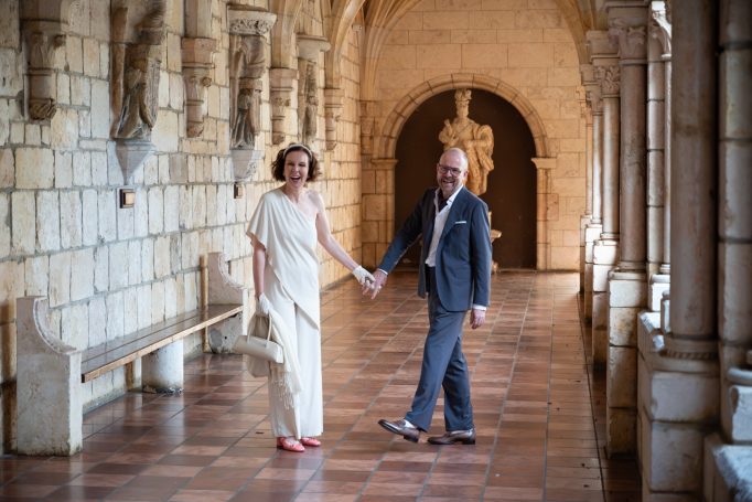 Couple holding hands and smiling in a historic stone corridor.