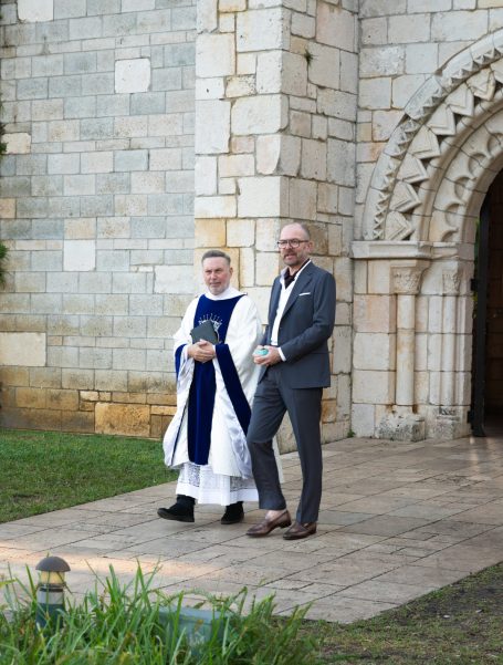 A clergy member in a white robe and a man in a suit walking outside a stone building.