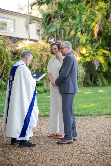 A couple exchanges vows during an outdoor wedding ceremony officiated by a priest.