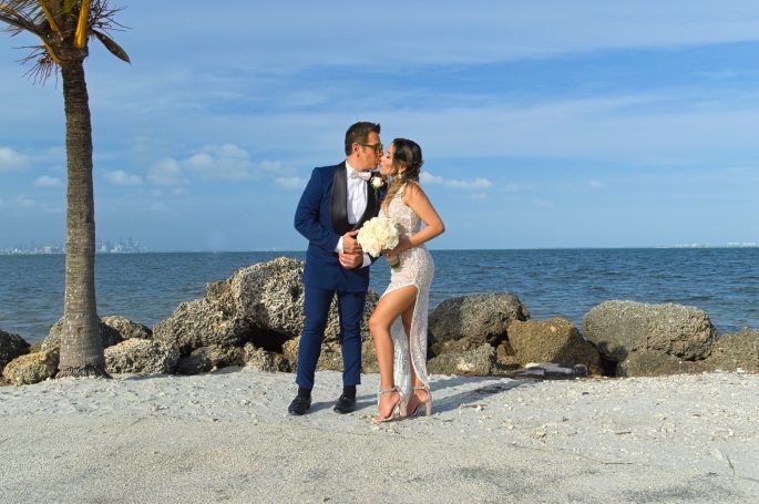 Couple embracing on a beach, with rocks and ocean in the background.