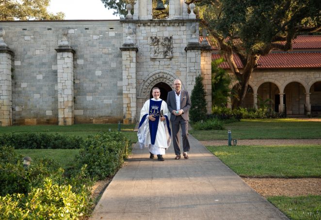 A person in religious attire walks alongside a man in formal clothing through a garden path.