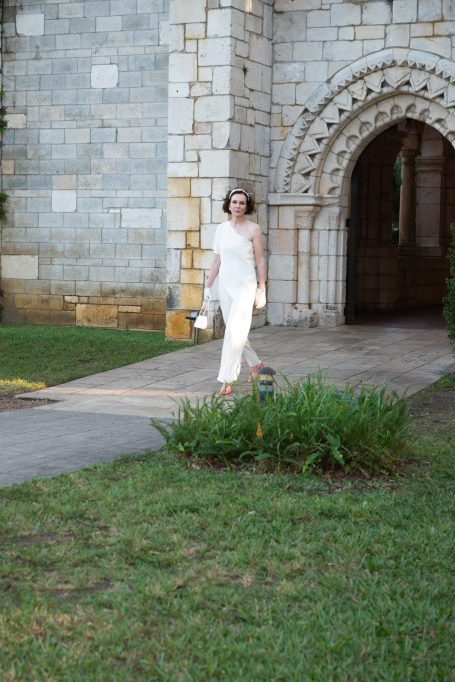 Woman in a white dress standing near a stone building and green grass.