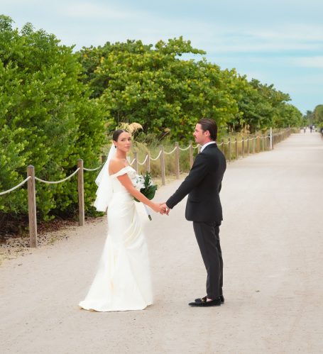Bride and groom holding hands on a sandy path surrounded by greenery.