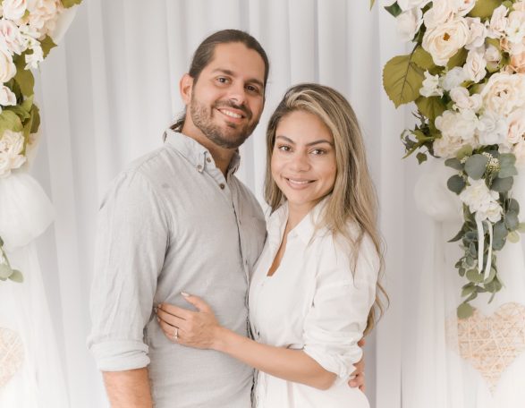 A couple smiles together in front of a decorative floral backdrop.