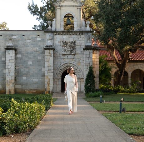 A person in light clothing walks along a path near a stone building and greenery.