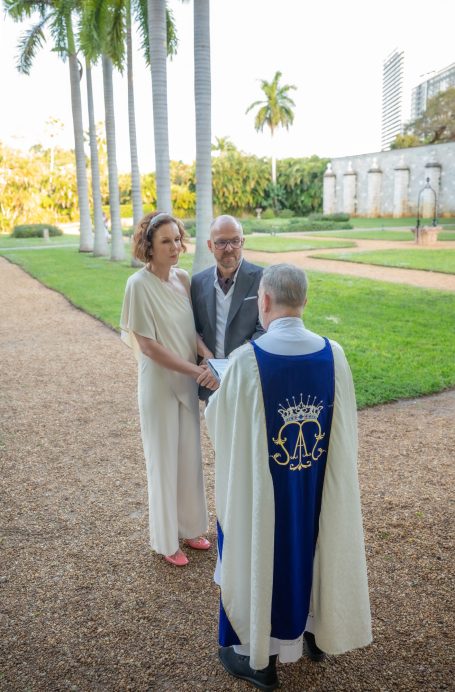 A couple stands before a officiant in a garden during a wedding ceremony. Palm trees in background.
