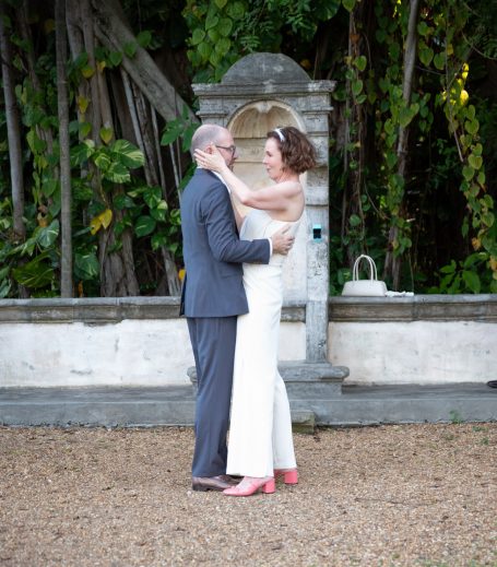 Bride and groom embrace outdoors, surrounded by greenery, on a gravel path.