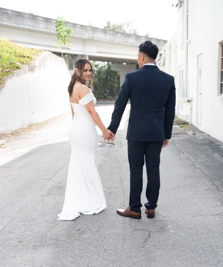 A bride and groom hold hands, walking down a sunlit alley in formal attire.