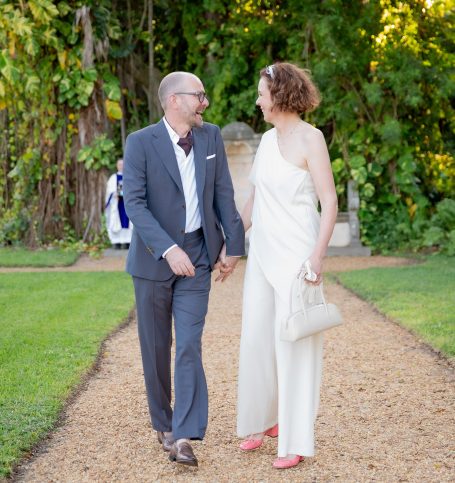 Couple holding hands, dressed elegantly, walking together on a garden pathway.