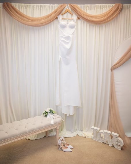 A white wedding dress hangs on a hanger with a floral bouquet and decorative drapery.
