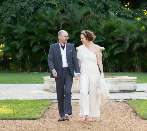 A couple walking hand in hand in a garden, dressed elegantly for a celebration.