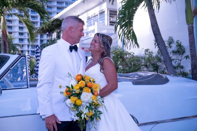 A smiling couple in formal attire, holding a bouquet, stands beside a vintage car.