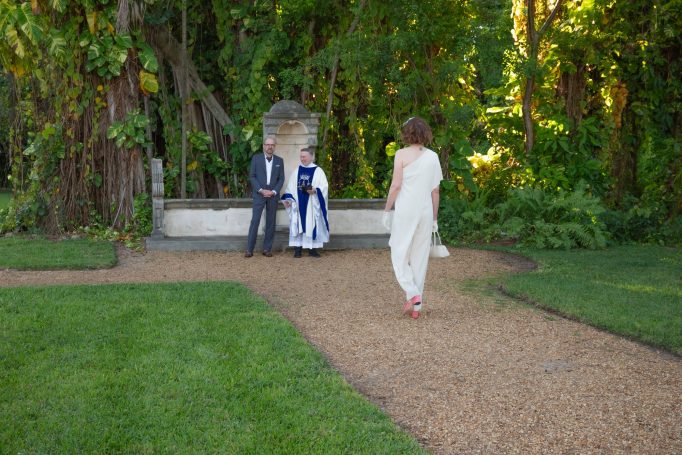 A woman in a white dress walks along a path toward seated individuals in a garden.