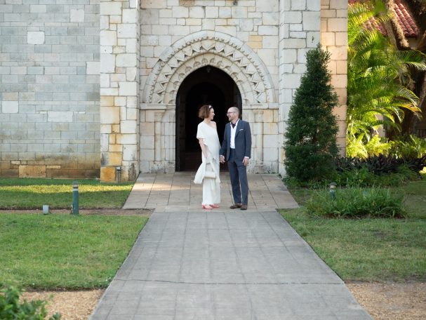 A man and woman walk together on a pathway near a stone building with an arched entrance.