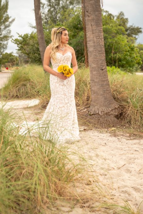 Bridal figure in a lace gown holding a bouquet, walking on a sandy path by palm trees.