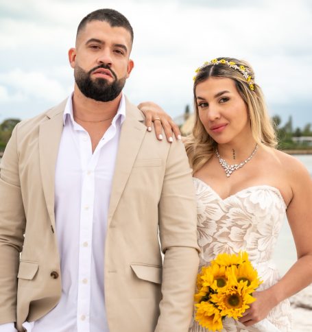 A bride and groom pose together, the bride holding sunflowers, near a body of water.