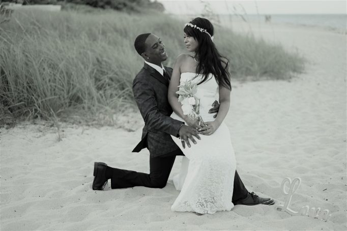 A couple poses romantically on the beach, with one kneeling and the other seated gracefully.