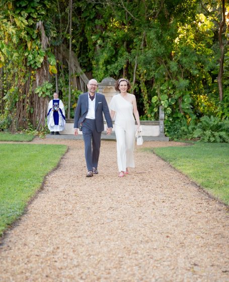 A couple walks hand in hand along a gravel path in a lush garden.