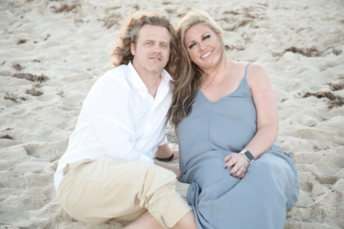 A couple sitting on the beach, smiling and dressed in casual attire.